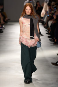 Model on a runway wearing a peach-pink polka-dot top with a ruffled hem, dark teal wide-leg pants, and a gray scarf with a black-and-white striped detail, carrying a blue bag.