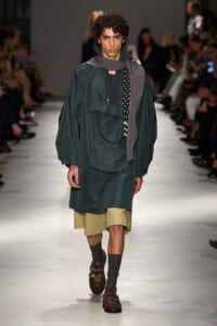 Male model walks a fashion runway wearing an oversized dark green coat over beige shorts, with a gray scarf and studded brown loafers.