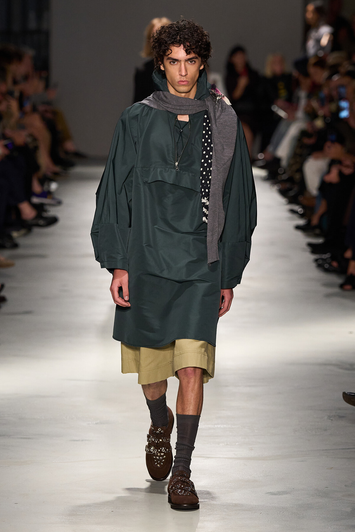 Male model walks a fashion runway wearing an oversized dark green coat over beige shorts, with a gray scarf and studded brown loafers.