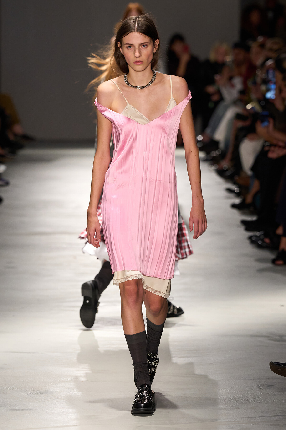 Models walks a pink satin slip dress with cream lace underlayer down a runway, pairing with dark socks and patent shoes.