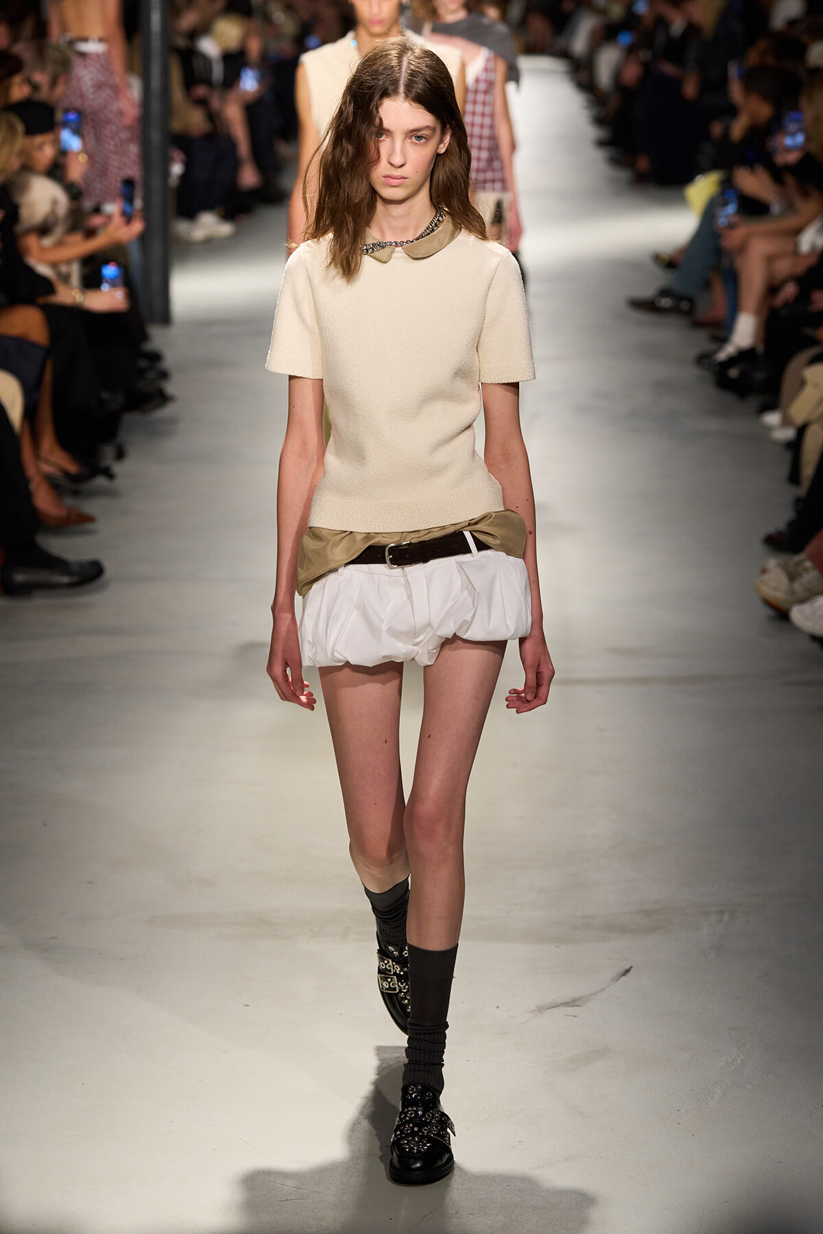 Model walks the runway wearing a cream short-sleeve knit top paired with white sculpted shorts and a belt; audience watches from both sides.