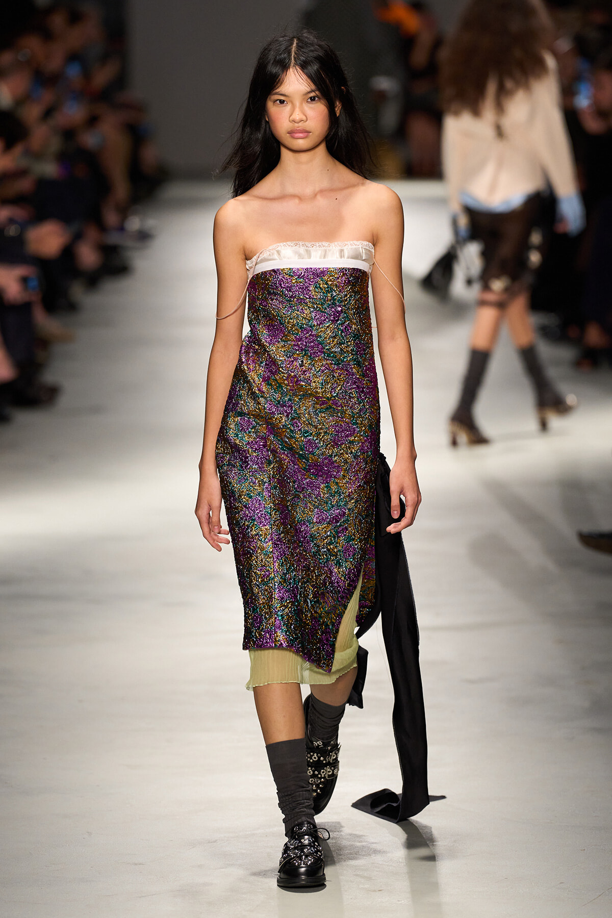 Model walking a runway in a strapless, sequined floral dress with purple, gold, and green tones, white neckline, and pale green underlayer; black boots with buckles and gray slouch socks.
