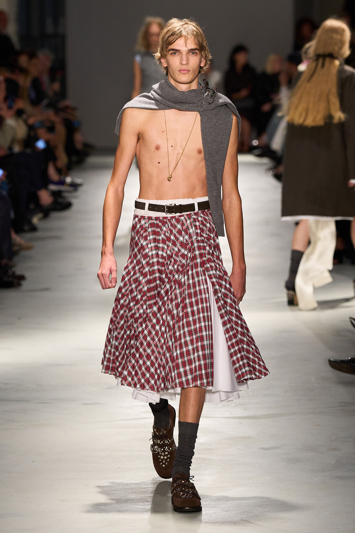 Male model on a fashion runway wearing a red plaid midi skirt, white underskirt, brown buckled boots, and a gray draped scarf with a pendant necklace.