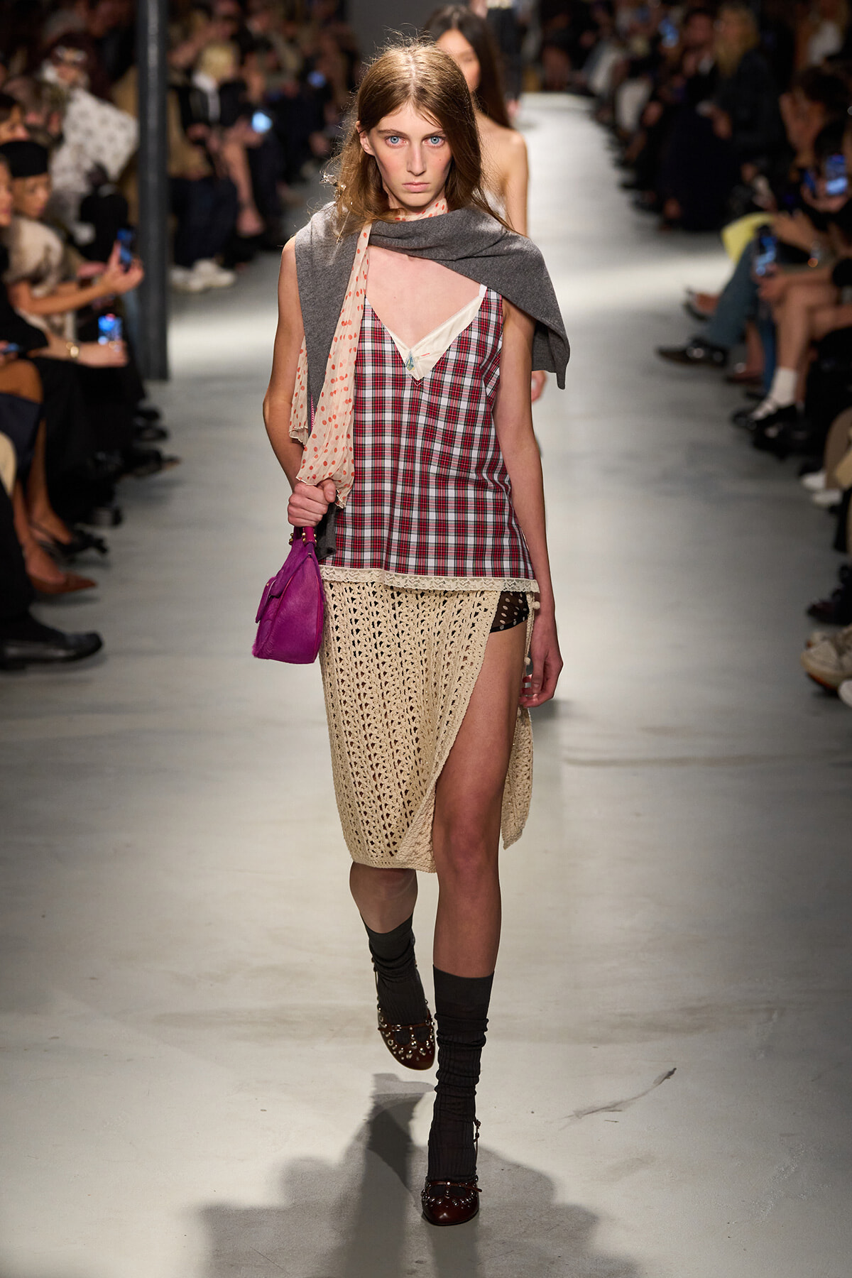 Model walking a fashion show in a plaid red-and-white top layered over a beige camisole, with a crochet beige skirt and a gray scarf draped over shoulders, carrying a purple handbag.