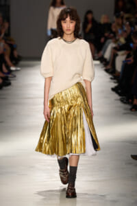 Model walks a runway in a cream knit sweater and a shiny gold pleated midi skirt, paired with dark socks and brown loafers.