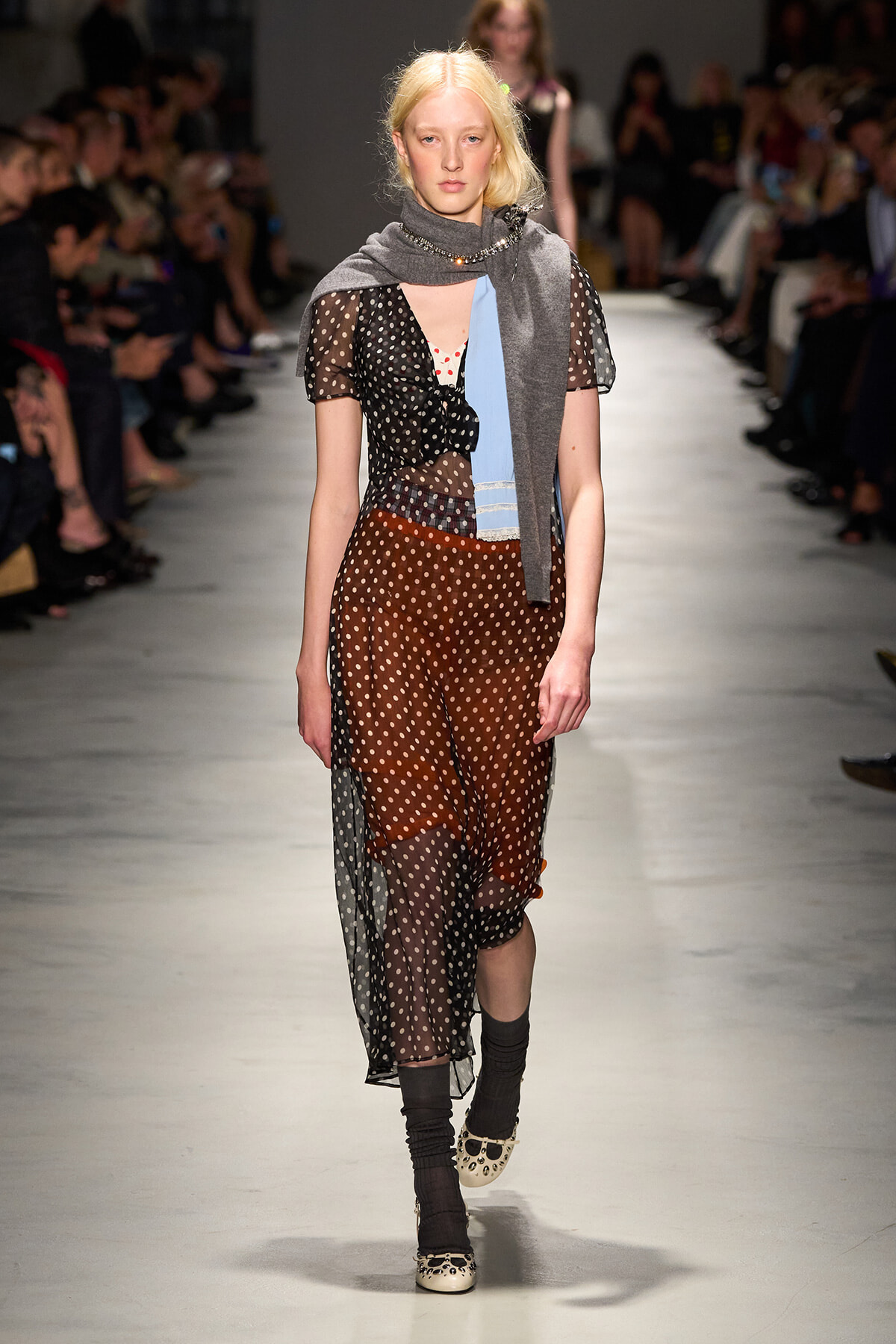 Model walks a fashion runway wearing a sheer black polka-dot dress over brown pants, with a gray scarf and light blue panel at the waist.