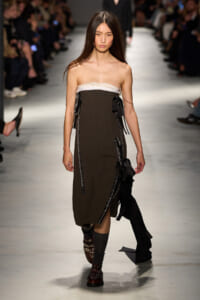 Model walks a fashion runway in a strapless brown dress with white neckline, black ribbons, and embellished boots.