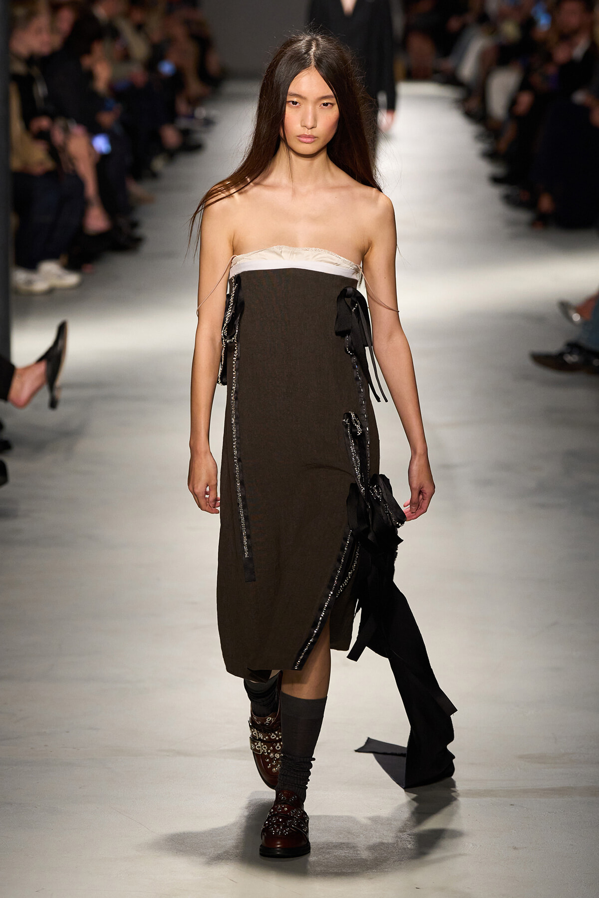 Model walks a fashion runway in a strapless brown dress with white neckline, black ribbons, and embellished boots.