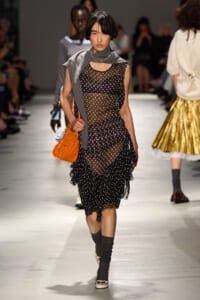 Model walking a runway in a sheer polka-dot dress with layered ruffles, gray scarf, and orange bag.