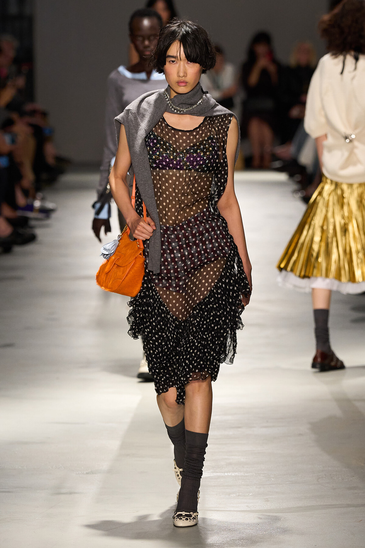Model walking a runway in a sheer polka-dot dress with layered ruffles, gray scarf, and orange bag.