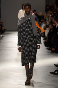 Model walking the runway in a black polka-dot coat with white cuffs, layered with gray scarf and orange sash, brown embellished loafers, and gray socks.