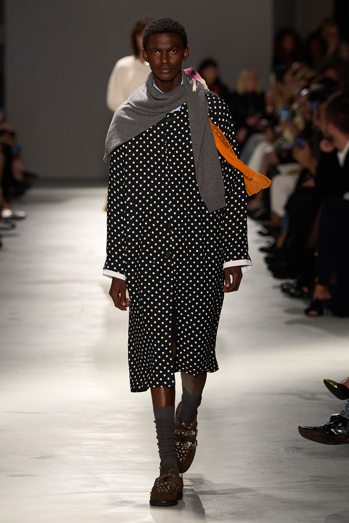Model walking the runway in a black polka-dot coat with white cuffs, layered with gray scarf and orange sash, brown embellished loafers, and gray socks.