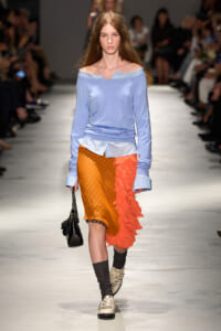 Model walks the runway in a pale blue off‑the‑shoulder top and a two‑tone orange skirt with ruffled petals, carrying a black handbag.