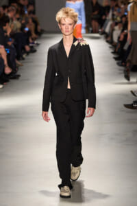 Model walking the runway in a black tailored suit with a floral shoulder embellishment, paired with matching trousers and embroidered cream loafers.