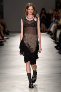 Model on a fashion runway wearing a sheer black mesh dress with a colorful sequined bustier, dotted underlay, and a black ruffled hem; audience blurred in background.