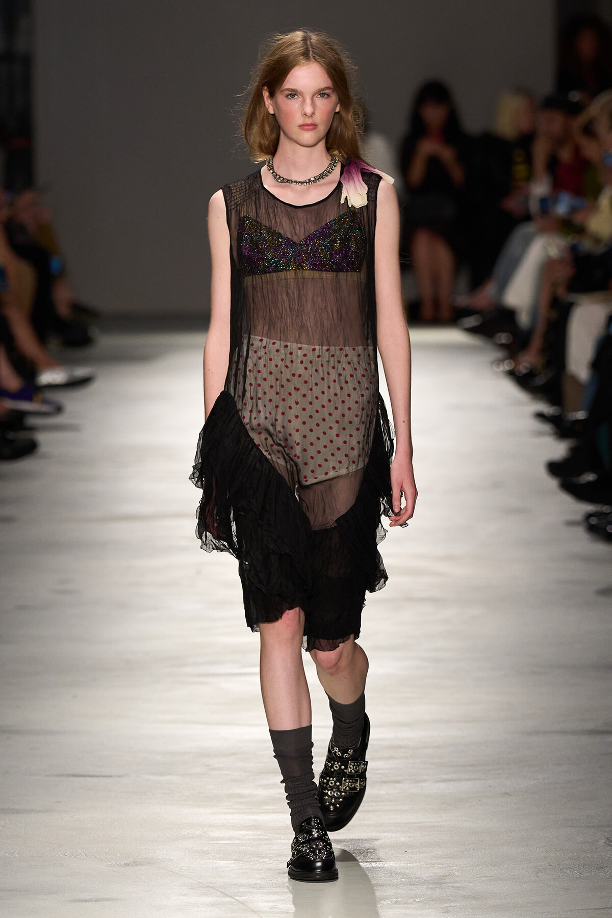 Model on a fashion runway wearing a sheer black mesh dress with a colorful sequined bustier, dotted underlay, and a black ruffled hem; audience blurred in background.