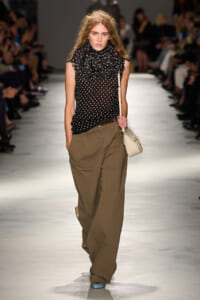 Model on a runway wearing a sheer black polka-dot blouse with a ruffled high neck and wide-legged khaki pants, carrying a cream shoulder bag.
