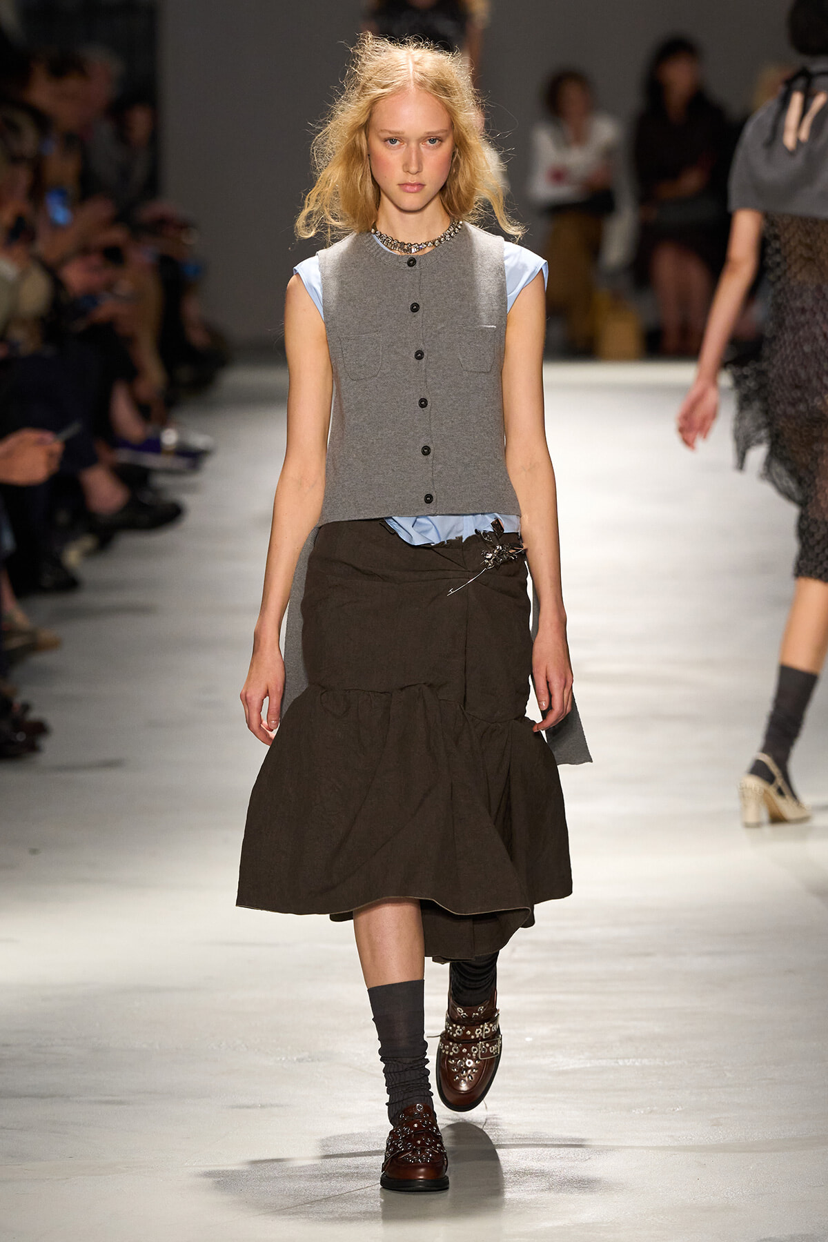Model walks the runway in a gray sleeveless buttoned vest over a light blue top and a brown tiered midi skirt.