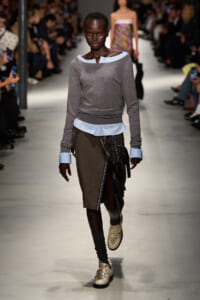 Front-view of a Black female model walking a fashion show runway in a layered gray knit top with light blue cuffs and hem, over a blue blouse, paired with a brown midi skirt with wrapped black fabric details and studs on beige lace-up shoes.