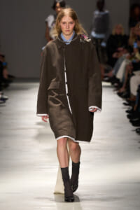 Model walking a runway in a loose brown coat over a white shirt, accessorized with a statement necklace and dark socks with flats.