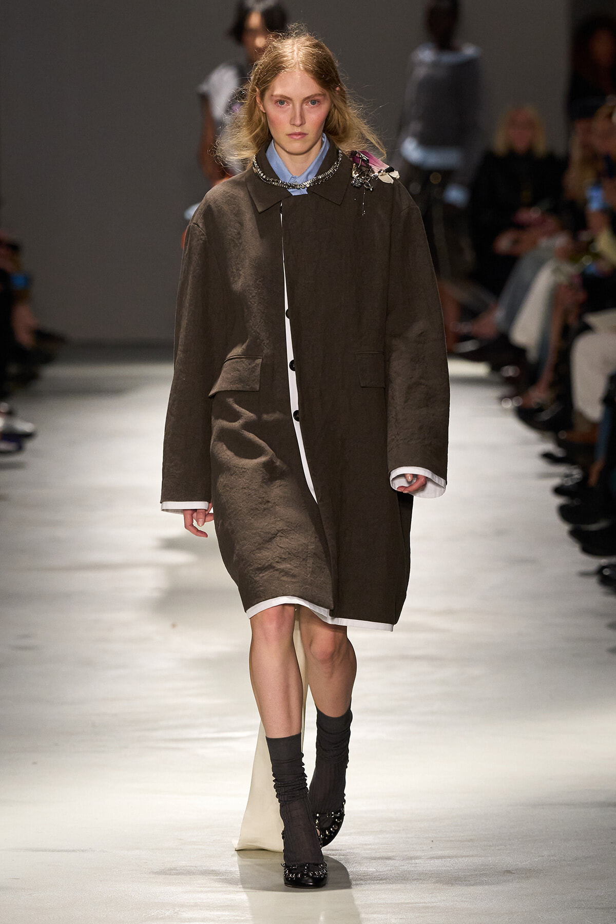 Model walking a runway in a loose brown coat over a white shirt, accessorized with a statement necklace and dark socks with flats.