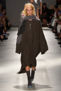 Model walking a runway in a loose brown tunic dress with a gray shawl and a black polka-dot scarf, paired with blue studded shoes.