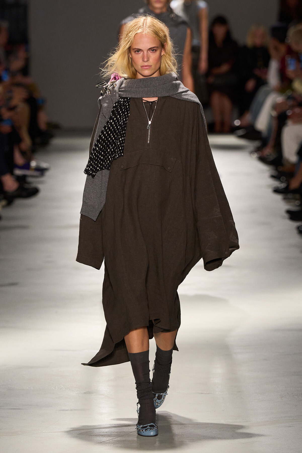 Model walking a runway in a loose brown tunic dress with a gray shawl and a black polka-dot scarf, paired with blue studded shoes.