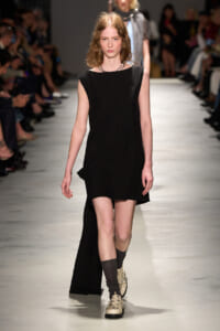 Model on a runway wearing a sleeveless black mini dress with a long back cape, beige lace-up shoes, and dark socks, with an audience in the background.