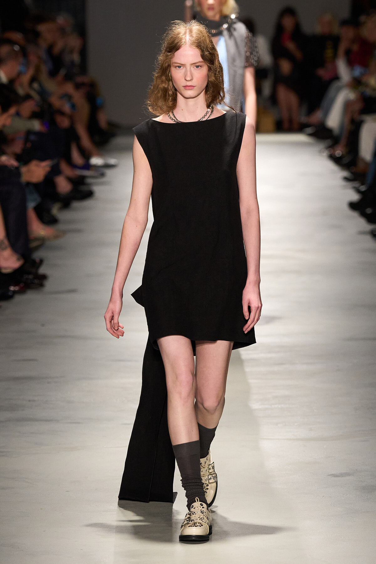 Model on a runway wearing a sleeveless black mini dress with a long back cape, beige lace-up shoes, and dark socks, with an audience in the background.
