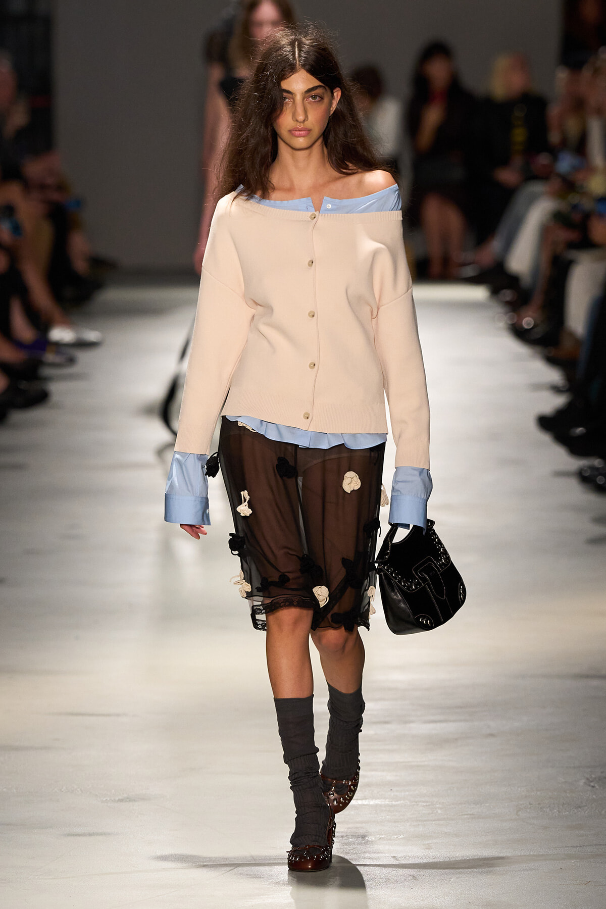 Model walking a runway in a pale pink off-shoulder cardigan over a blue blouse, paired with a sheer black floral-embellished skirt, gray socks, brown shoes, and a black handbag.