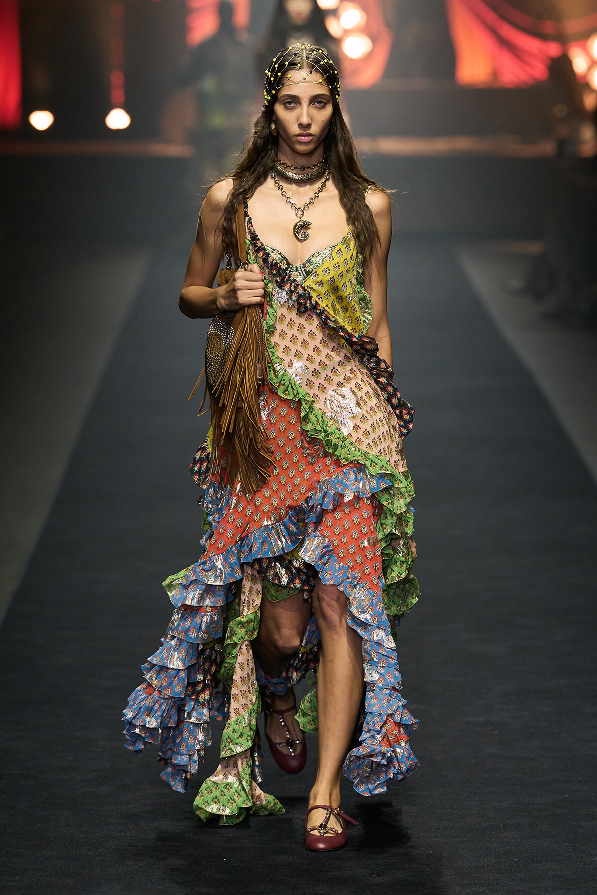 Model walks a runway wearing a multi‑colored patchwork, ruffled boho dress with fringe bag and burgundy flats.