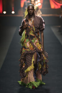 Model walking a fashion runway in a layered, colorful printed dress with a brown bomber jacket and green fringe bag accessory.