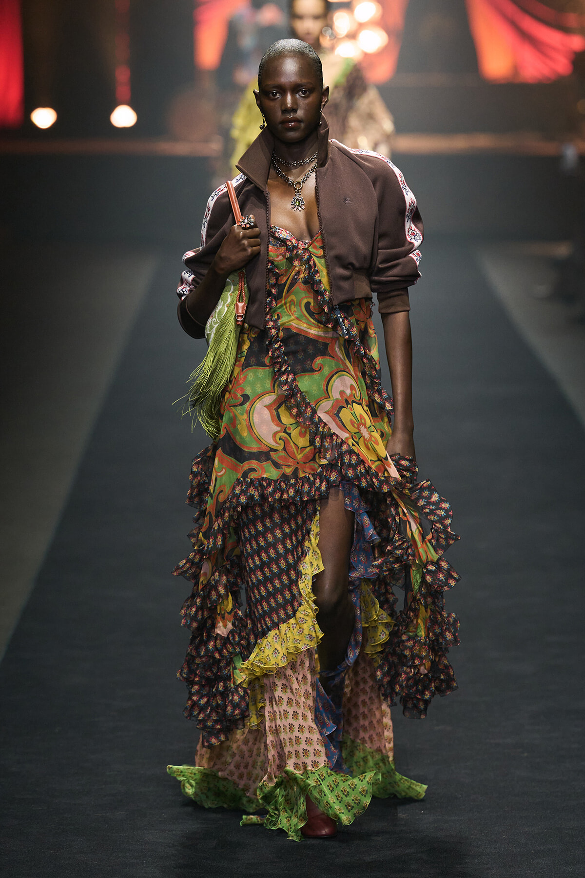 Model walking a fashion runway in a layered, colorful printed dress with a brown bomber jacket and green fringe bag accessory.