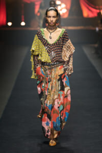 Model walking a runway in a colorful patchwork boho outfit with layered ruffle sleeves and wide belt, gold jewelry, and patterned pants.