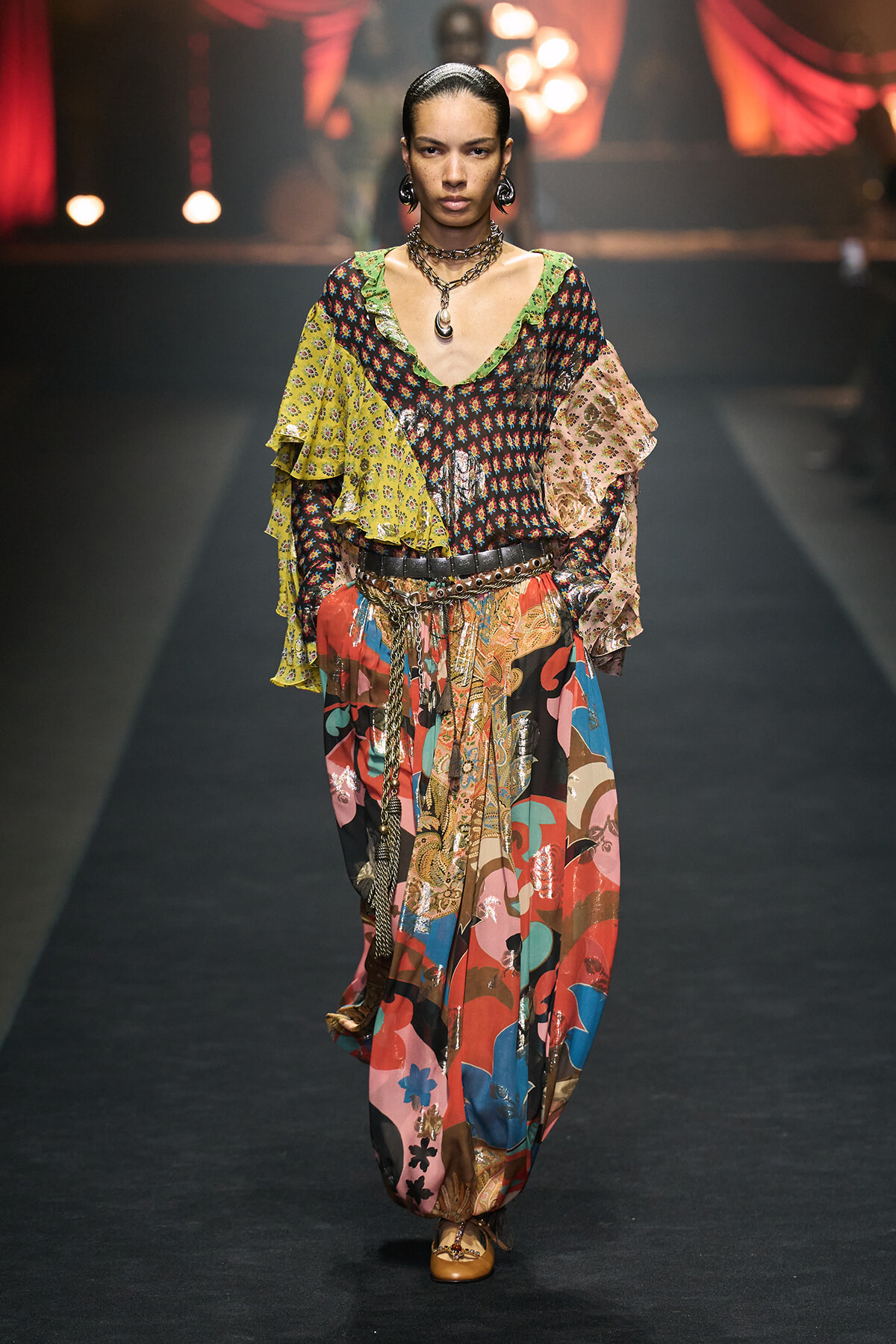 Model walking a runway in a colorful patchwork boho outfit with layered ruffle sleeves and wide belt, gold jewelry, and patterned pants.