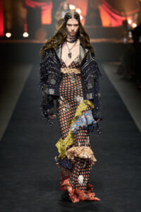 Model walking the runway in a patchwork floral maxi dress with layered ruffles and a black studded jacket, accessorized with chains and a beaded headpiece.