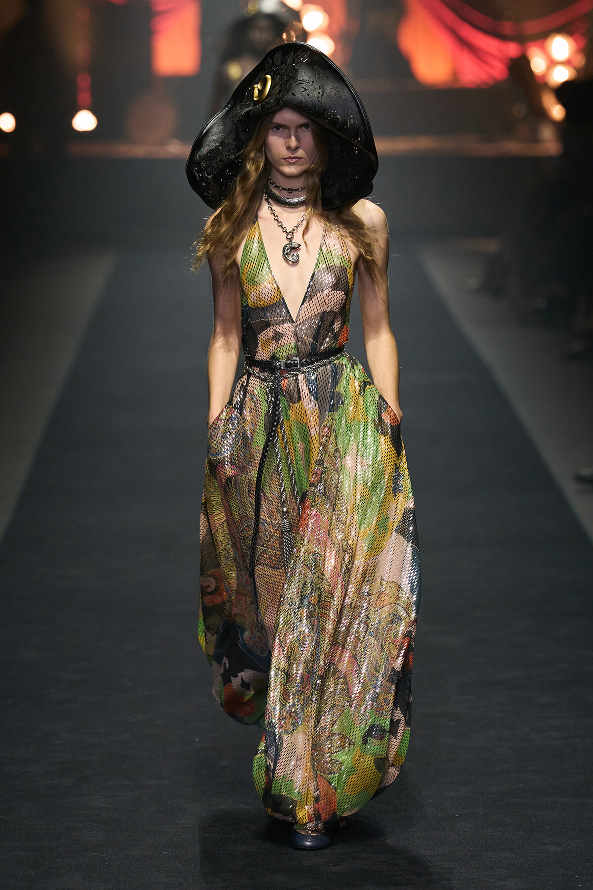 Model on runway wearing a colorful, patterned, flowing dress with a deep V-neck and multiple necklaces, topped by a large black ornate hat.
