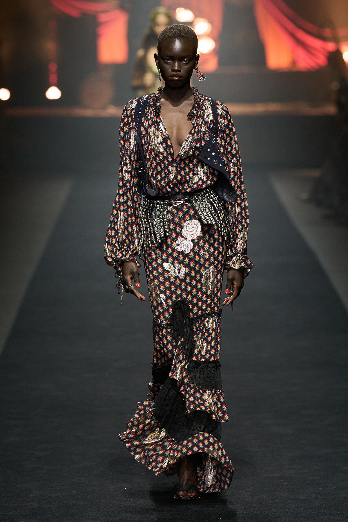 Model walking a runway in a dark floral maxi dress with ruffles and a metallic belt.