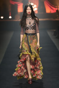 Model walking a fashion runway in a sheer floral top and multi-layered, ruffled skirt with bold jewelry and maroon shoes.
