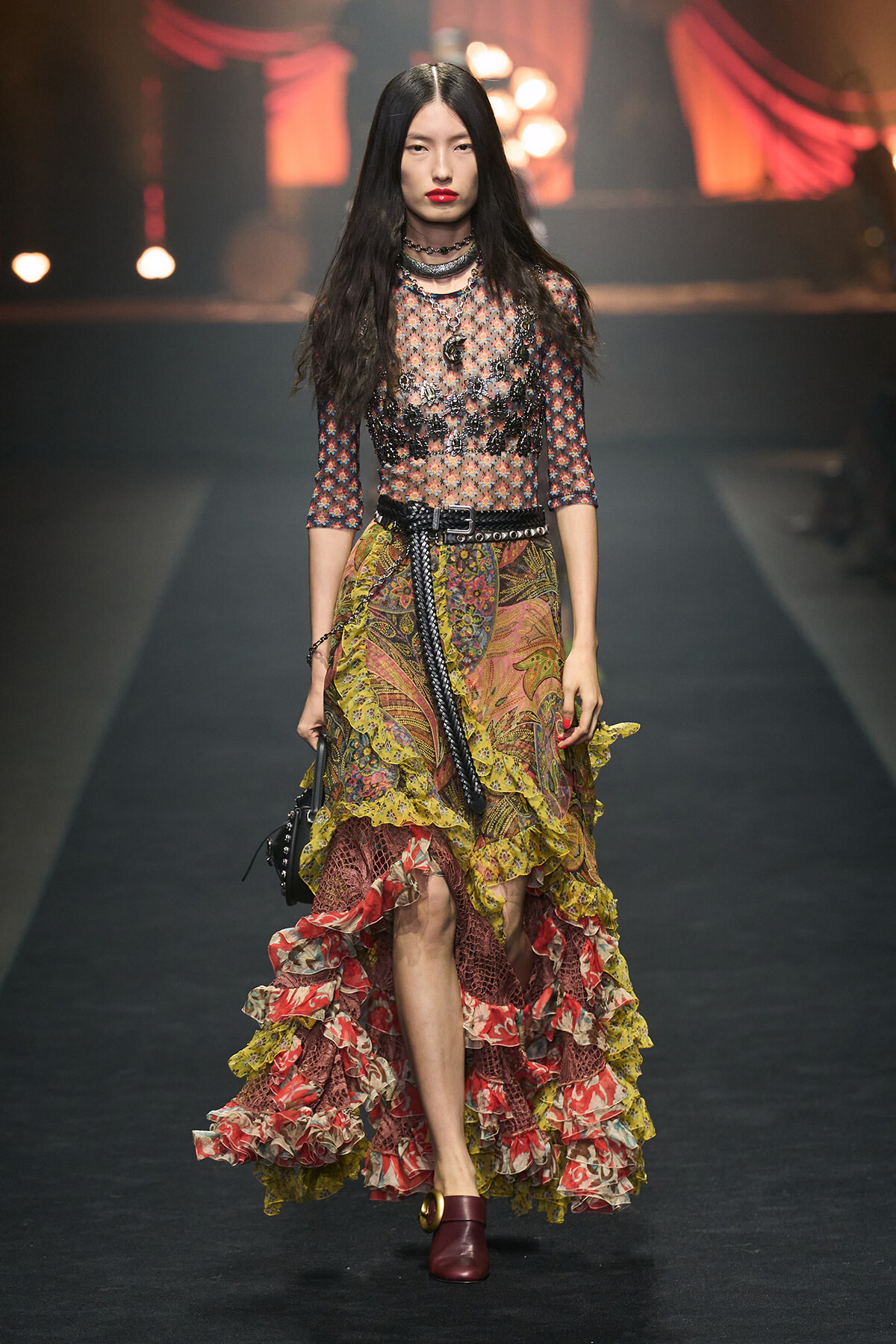 Model walking a fashion runway in a sheer floral top and multi-layered, ruffled skirt with bold jewelry and maroon shoes.