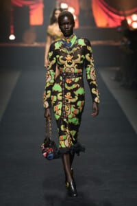 Model walks runway in a long black dress with bright green, orange, and yellow baroque pattern, cinched with a studded belt, carrying a small patterned bag.