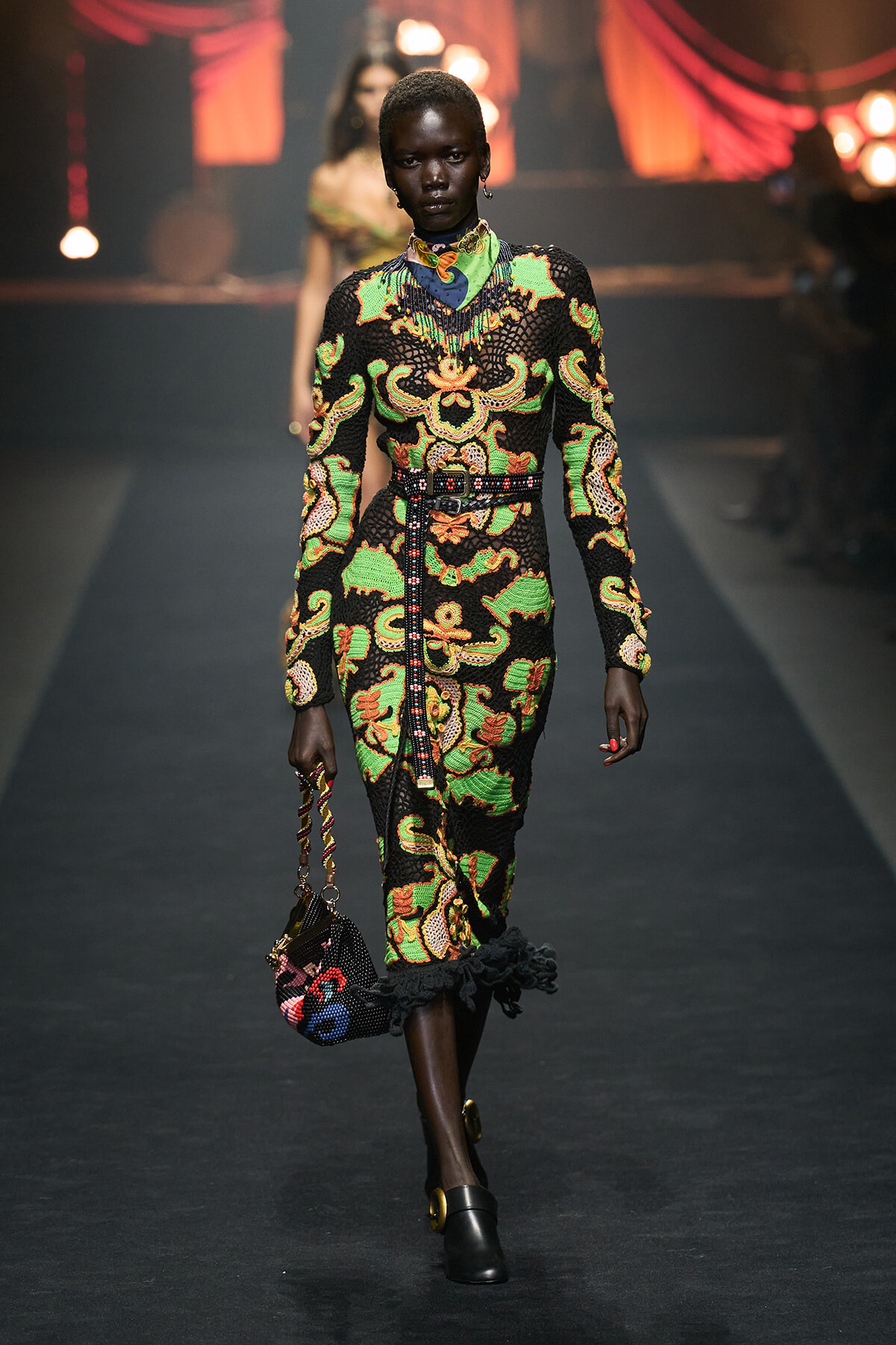 Model walks runway in a long black dress with bright green, orange, and yellow baroque pattern, cinched with a studded belt, carrying a small patterned bag.