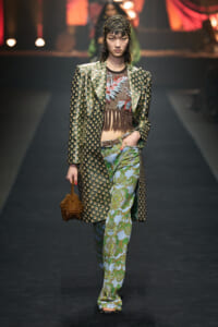 Model walking a runway in a green patterned long coat, fringe mesh top, and paisley-print flared pants, holding a small brown handbag.