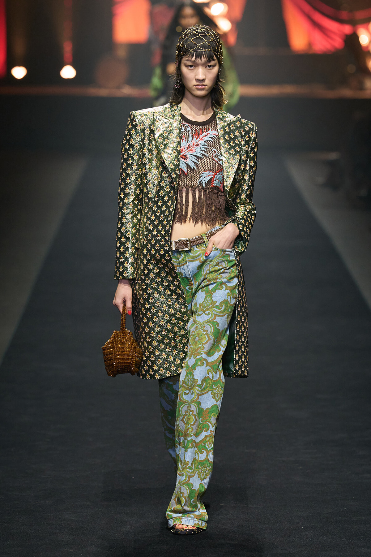 Model walking a runway in a green patterned long coat, fringe mesh top, and paisley-print flared pants, holding a small brown handbag.