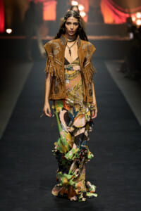Model walking a runway in a brown fringed suede vest with beadwork over a colorful ruffled dress, accessories, and headpiece.