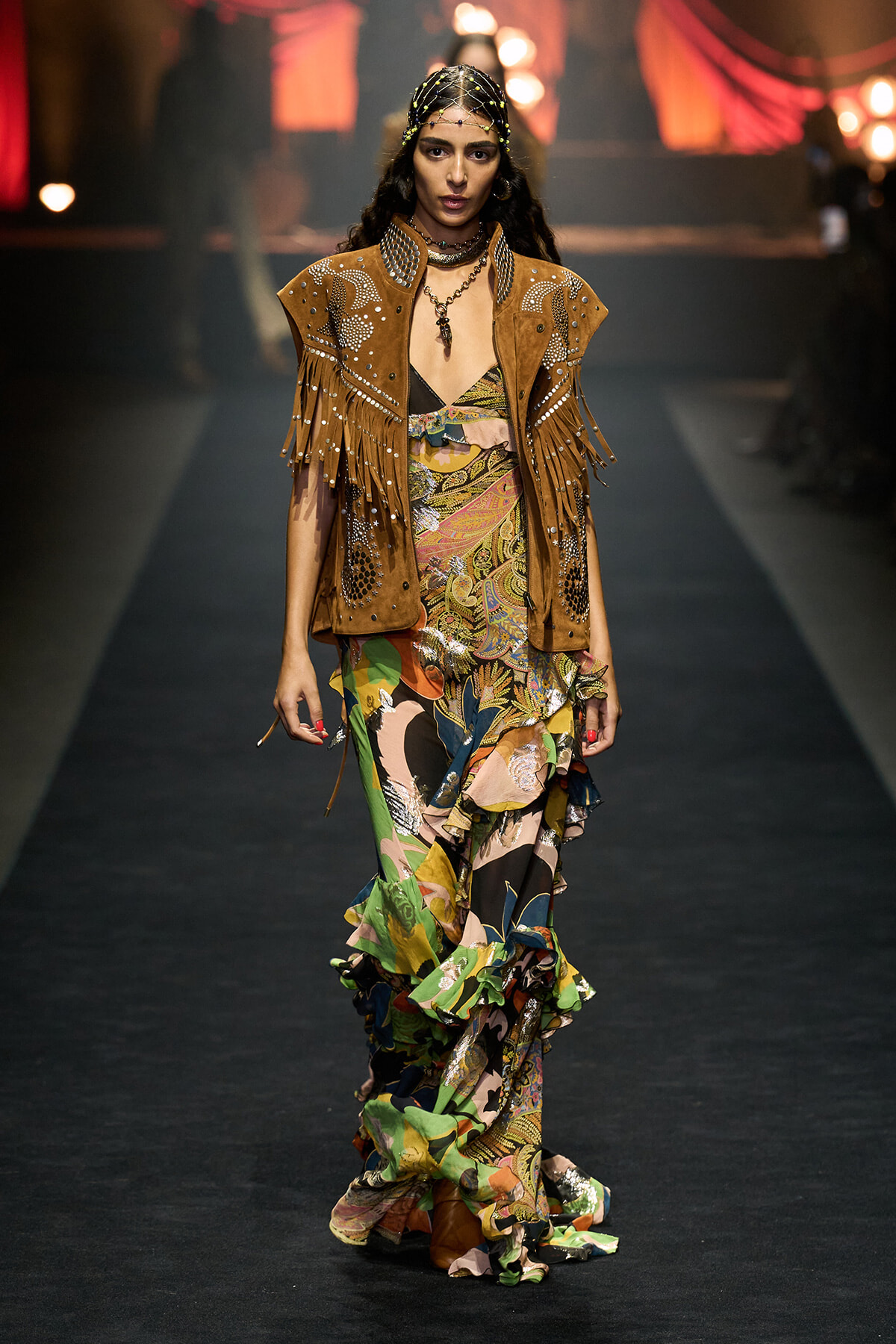 Model walking a runway in a brown fringed suede vest with beadwork over a colorful ruffled dress, accessories, and headpiece.