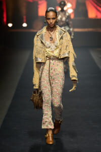 Model walks the runway in a beige fringed jacket with studs over floral lace trousers, brown boots, and bold jewelry.