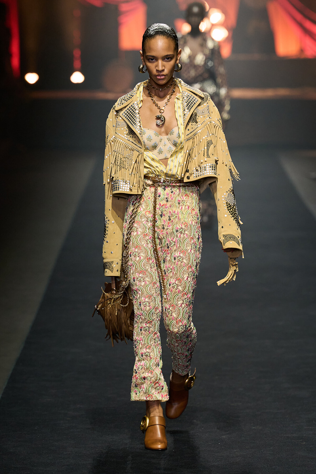 Model walks the runway in a beige fringed jacket with studs over floral lace trousers, brown boots, and bold jewelry.