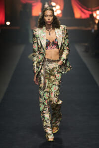 Model on a fashion show runway wearing a metallic floral suit with a matching pants, a pink patterned bra top, belt, and headpiece.