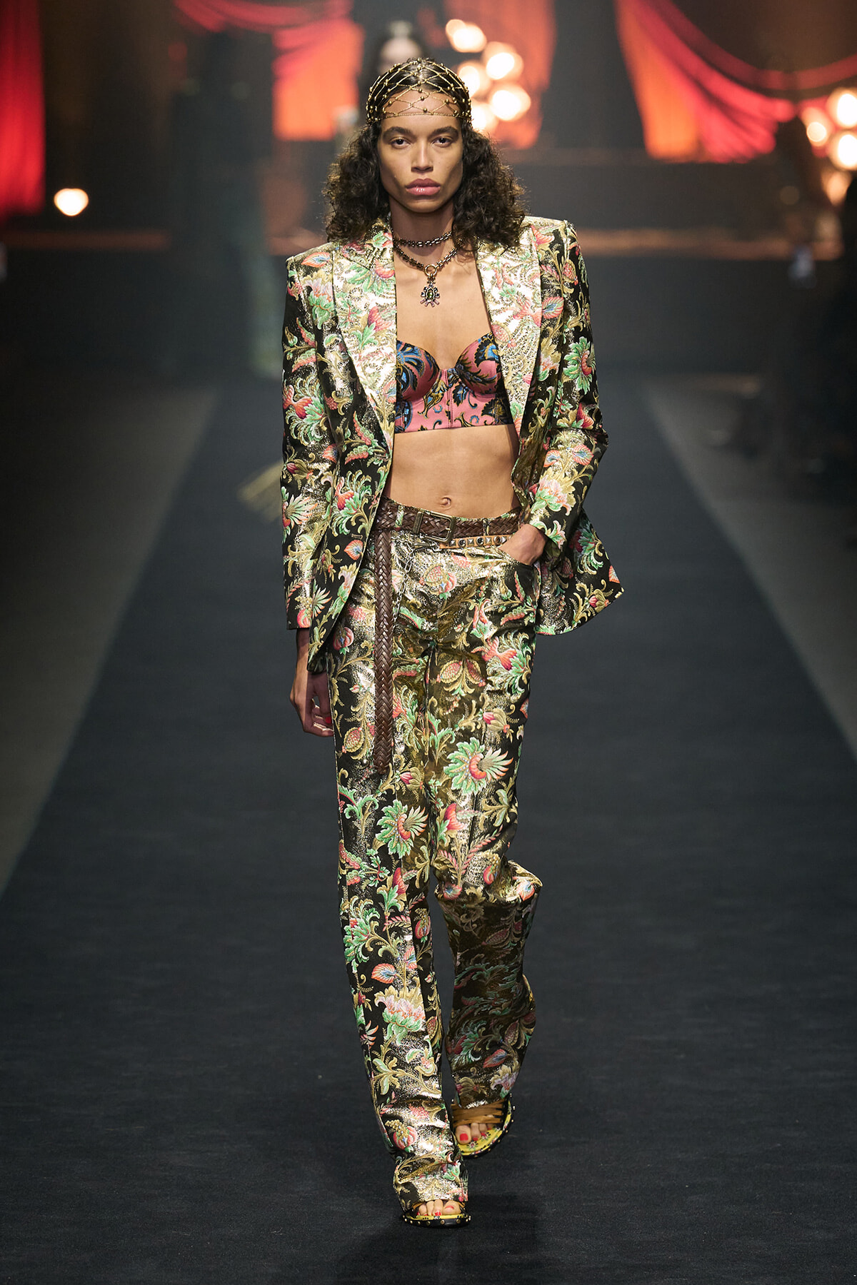 Model on a fashion show runway wearing a metallic floral suit with a matching pants, a pink patterned bra top, belt, and headpiece.